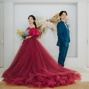 ドライフラワーの前に離れて立つスーツの新郎とロングトレーンのボルドーのカラードレスの花嫁。洋装スタジオフォト。