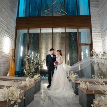 青いガラス窓の祭壇の前でベールをなびかせて寄り添う、新郎新婦を引きで写したチャペルフォト