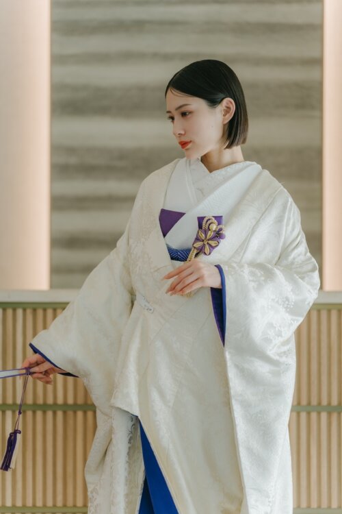 木目のカウンターがあるスタジオで、紫の筥迫と扇子を手に視線を落とす白無垢の花嫁。青い掛下が映えるスタジオフォトの一枚