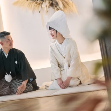 綿帽子の白無垢の花嫁と紋付袴の新郎が正座して見つめ合うワンシーン。前ボケを生かした和装スタジオフォト。