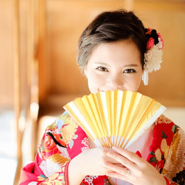 金色の扇子で口元を隠しながら微笑む新婦、赤い振袖と白と赤の花飾り。