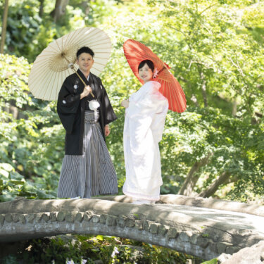 石橋の上で白い和傘と赤い和傘を差して並ぶ黒紋付袴の新郎と白無垢の花嫁。日本庭園で撮る和装前撮り