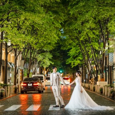 夜の横断歩道を車のライトの中で渡るベージュのスーツの新郎とウェディングドレスの花嫁。丸の内の街中で撮る洋装のナイトフォト前撮り