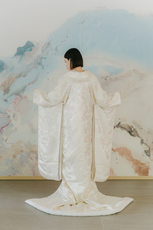 淡いブルーの壁画の前で両袖を広げ、後ろ姿を見せる白無垢の花嫁。背中から裾まで続く地紋が分かる和装フォトウェディングのバックスタイル