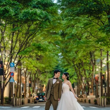 街路樹の並ぶ通りの真ん中で額を寄せ合うオリーブのスーツの新郎とウェディングドレスの花嫁を引きでとらえた一枚。丸の内の街中で撮る洋装ロケーションフォト