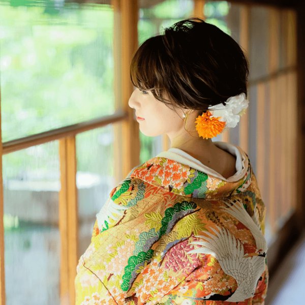 華やかな鶴の刺繍が入った色打掛姿の新婦の後ろ姿、オレンジと白の生花イヤリング、窓辺で外を眺める横顔。
