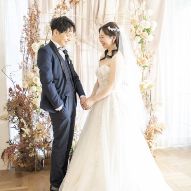 ドライフラワーのアーチの前で手をつないで見つめ合う、ネイビーのタキシードの新郎とウェディングドレスの花嫁のスタジオフォト