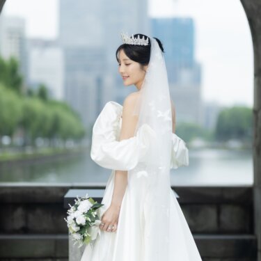 石のアーチ越しにお堀と高層ビルを望み、ブーケを持って立つティアラのウェディングドレスの花嫁の後ろ姿。丸の内で撮る洋装前撮り