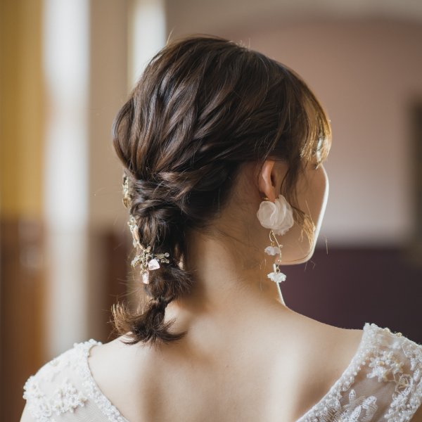 レース刺繍のドレスを着た新婦の三つ編みヘアスタイル、白い花のイヤリングとビジューの髪飾り。