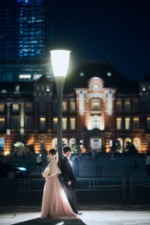 ライトアップされた東京駅を背に、街灯のそばで寄り添う新郎とピンククリームのカラードレスの花嫁。夜の洋装前撮り。