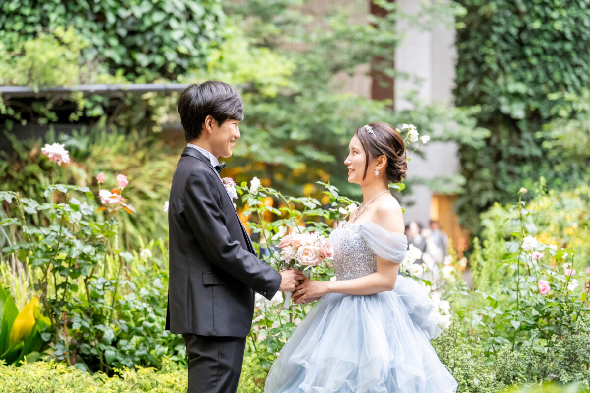 三菱一号館前のバラ園でブーケを手に向き合う、タキシードの新郎とブルーのカラードレスの花嫁の前撮り