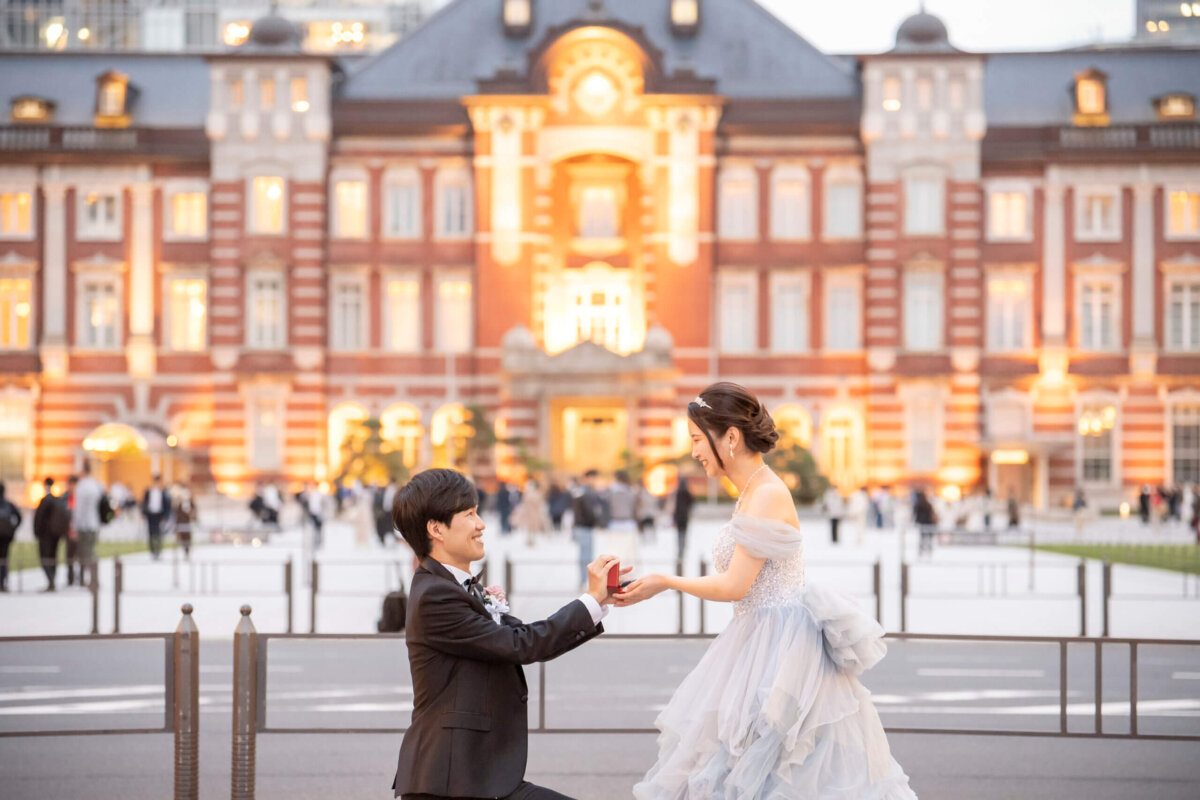 赤レンガの東京駅丸の内駅舎前の広場で片膝をつき、ブルーのカラードレスの花嫁に指輪を差し出すプロポーズの前撮り