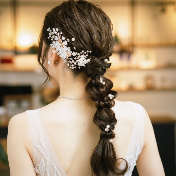 パールとフラワーモチーフの華やかなヘアアクセサリーを編み込んだ三つ編みヘアスタイル、白いドレスの後ろ姿。