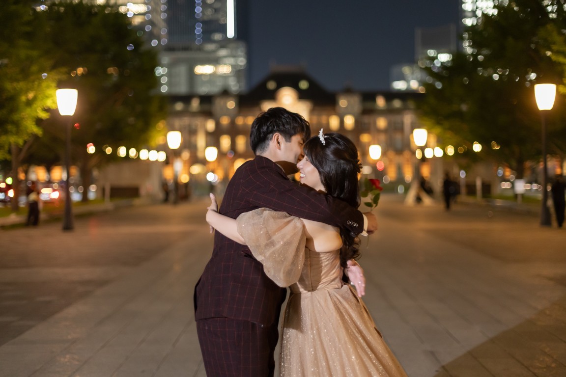 ライトアップされた東京駅丸の内駅舎の前で赤いバラを持って抱き合うボルドーのスーツの新郎とベージュのカラードレスの花嫁。東京駅で撮る洋装前撮りのナイトフォト