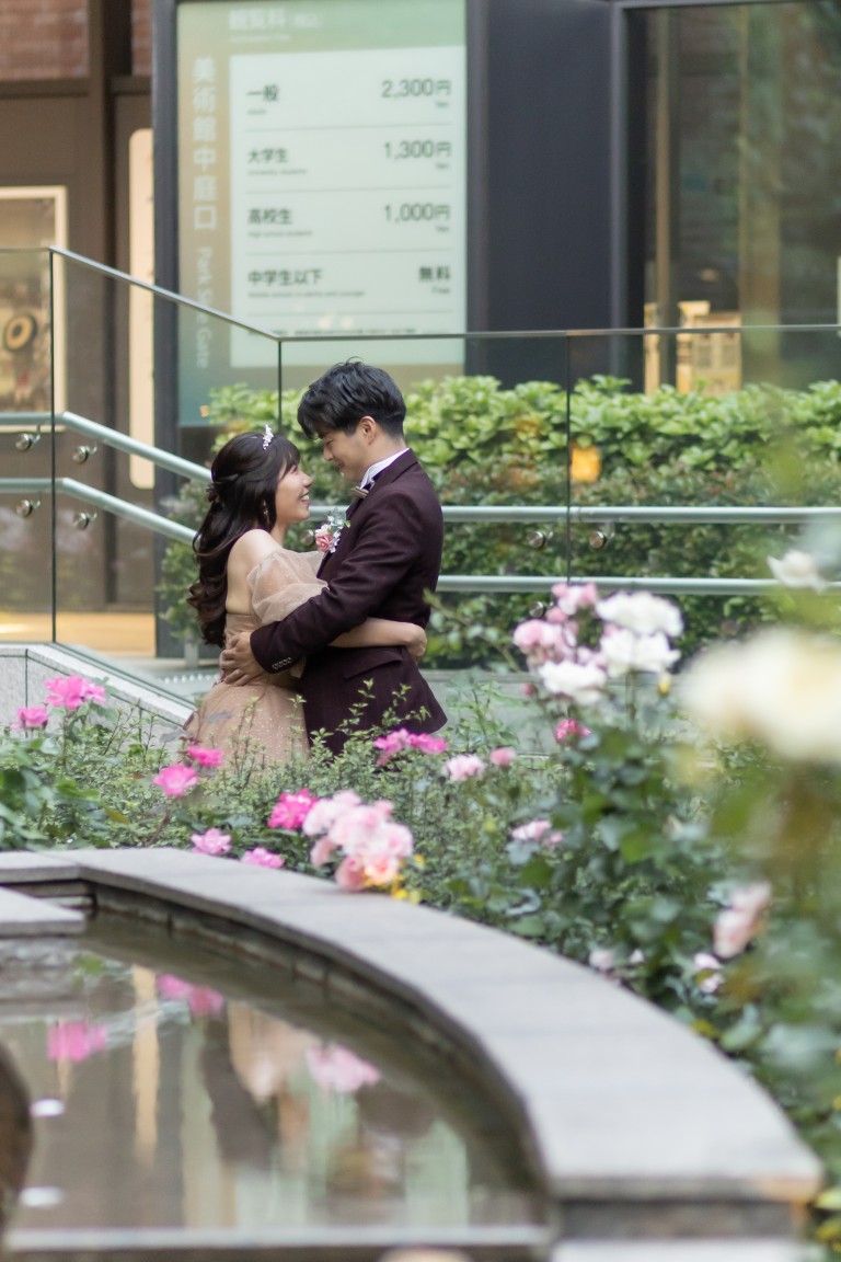 バラの咲く花壇と水盤のそばで抱き合うボルドーのスーツの新郎とベージュのカラードレスの花嫁を縦位置でとらえた一枚。丸の内のバラ園で撮る洋装前撮り