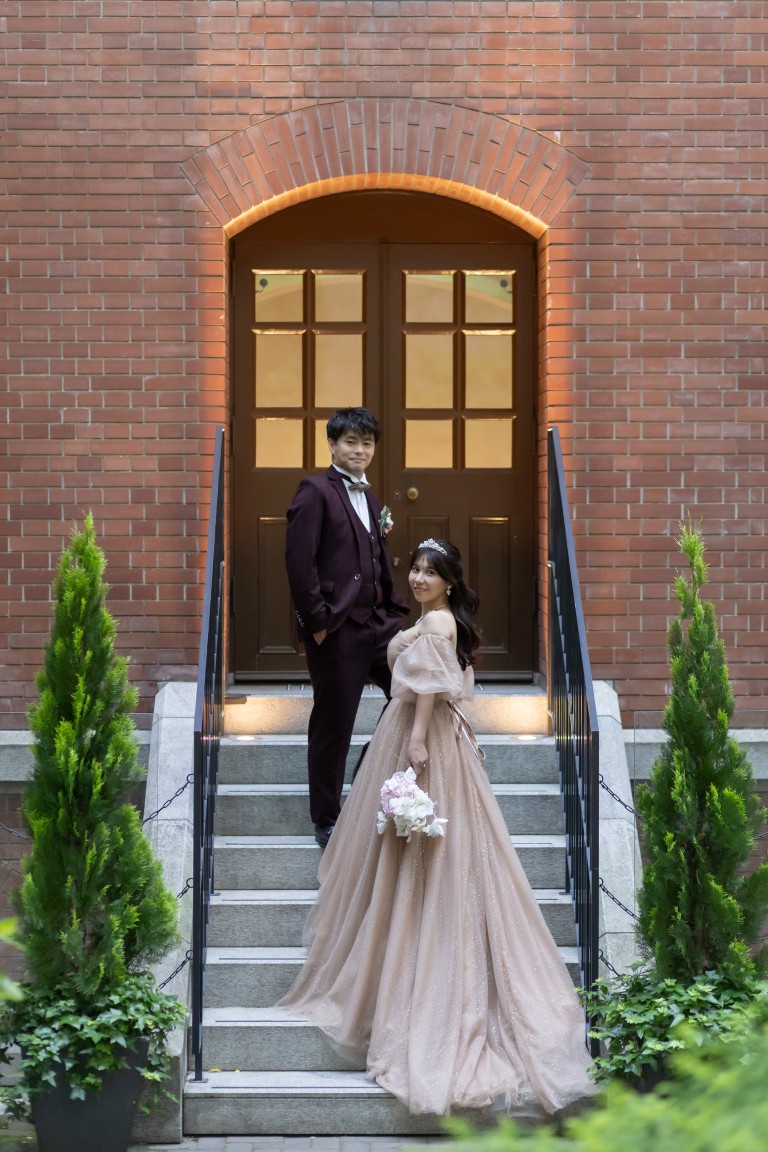 赤レンガのアーチの扉の階段に並ぶボルドーのスーツの新郎とベージュのカラードレスの花嫁を縦位置でとらえた一枚。丸の内で撮る洋装前撮り