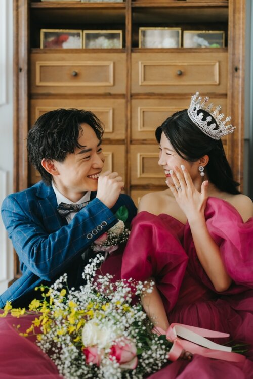 木の棚の前でブーケを持って笑い合うネイビーのスーツの新郎とティアラのピンクのカラードレスの花嫁を縦位置でとらえた一枚。東京駅店のスタジオで撮る洋装前撮り