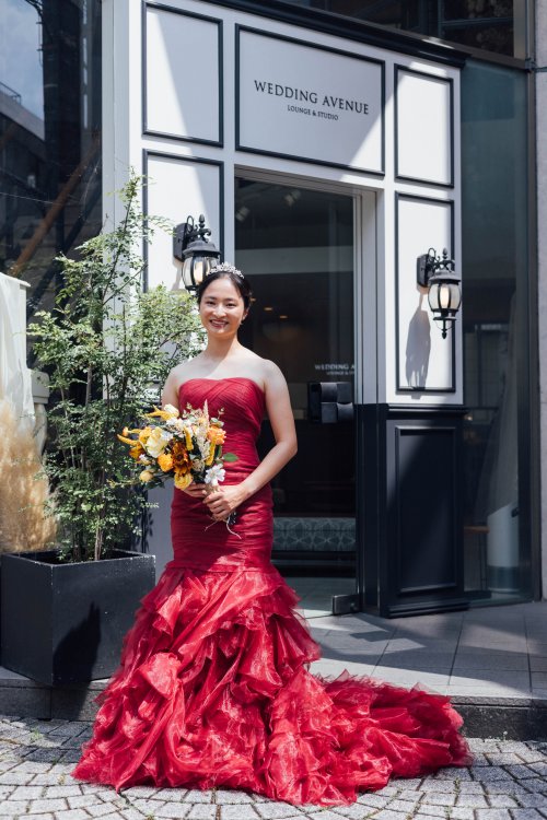 『WEDDING AVENUE』の看板のある店舗の前でブーケを持つ赤いマーメイドドレスの花嫁を縦位置でとらえた一枚。東京駅店の前で撮る洋装前撮りのカラードレスカット