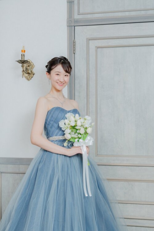 白い扉の前で白いブーケを持つくすみブルーのカラードレスの花嫁を縦位置でとらえた一枚。東京駅店のスタジオで撮る洋装前撮りのカラードレスカット