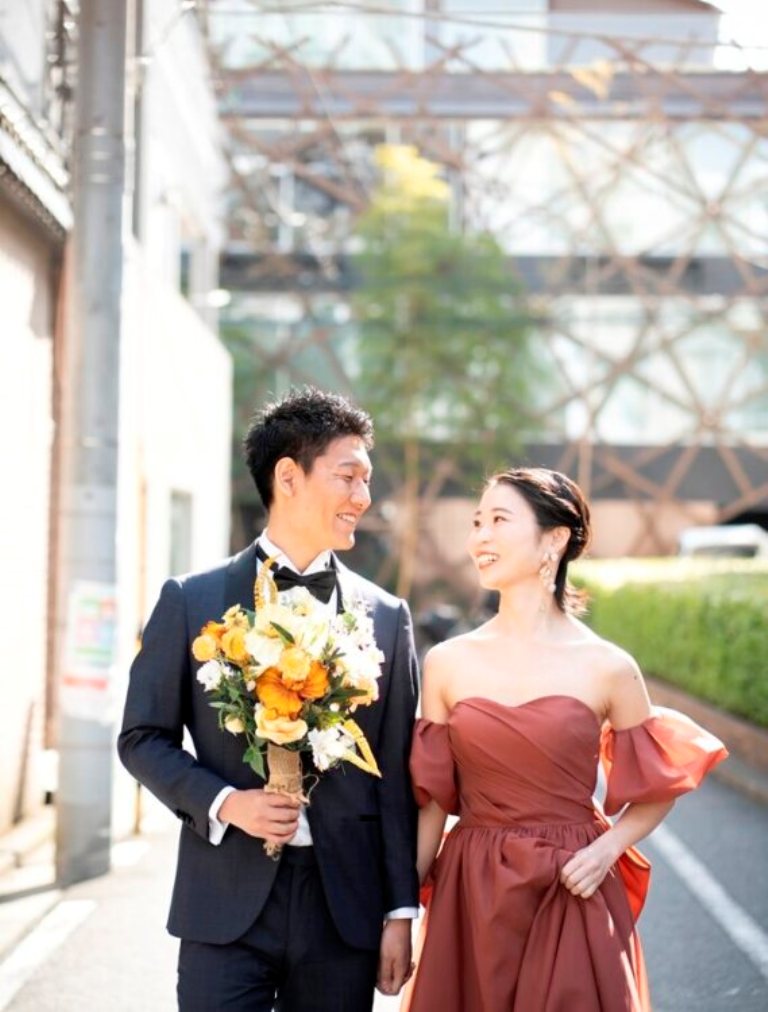 路地でオレンジのブーケを持って並ぶネイビーのタキシードの新郎とオレンジのカラードレスの花嫁を縦位置でとらえた一枚。東京駅店の前で撮る洋装前撮り