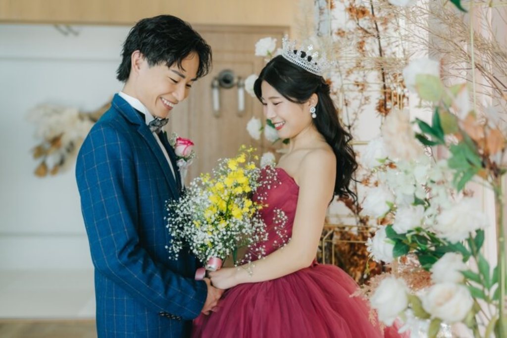 ドライフラワーの前で花嫁の手を取るネイビーのスーツの新郎とブーケを持つティアラの赤いカラードレスの花嫁。東京駅店のスタジオで撮る洋装前撮り