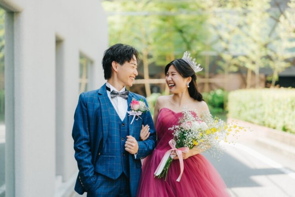 緑を背に腕を組んで笑い合うネイビーのスーツの新郎とブーケを持つティアラの赤いカラードレスの花嫁。東京駅店の前で撮る洋装前撮り