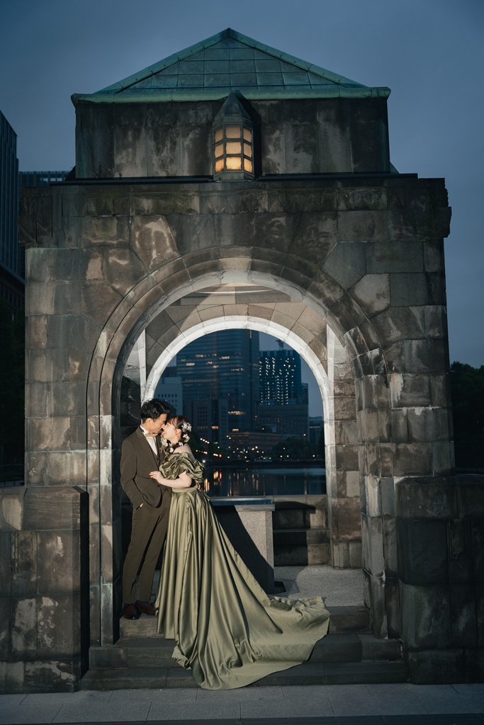 ライトアップされた石のアーチの門の下でキスをするオリーブのスーツの新郎とオリーブのカラードレスの花嫁を縦位置で引きでとらえた一枚。丸の内で撮るナイトフォト前撮り