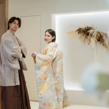 ドライフラワーを飾ったスタジオで、グレージュの紋付袴の新郎と淡い色打掛を羽織って振り返る花嫁の和装スタジオフォト